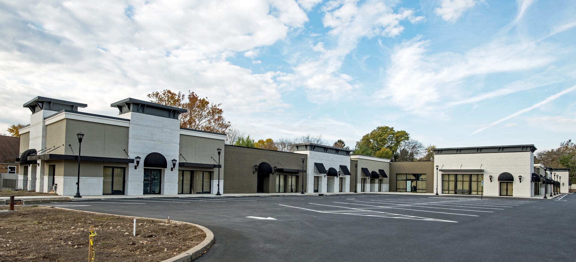 A newly completed multi-tenant retail center with stucco and masonry storefronts, black awnings, and carriage lamps, fronted by a freshly paved and striped parking lot