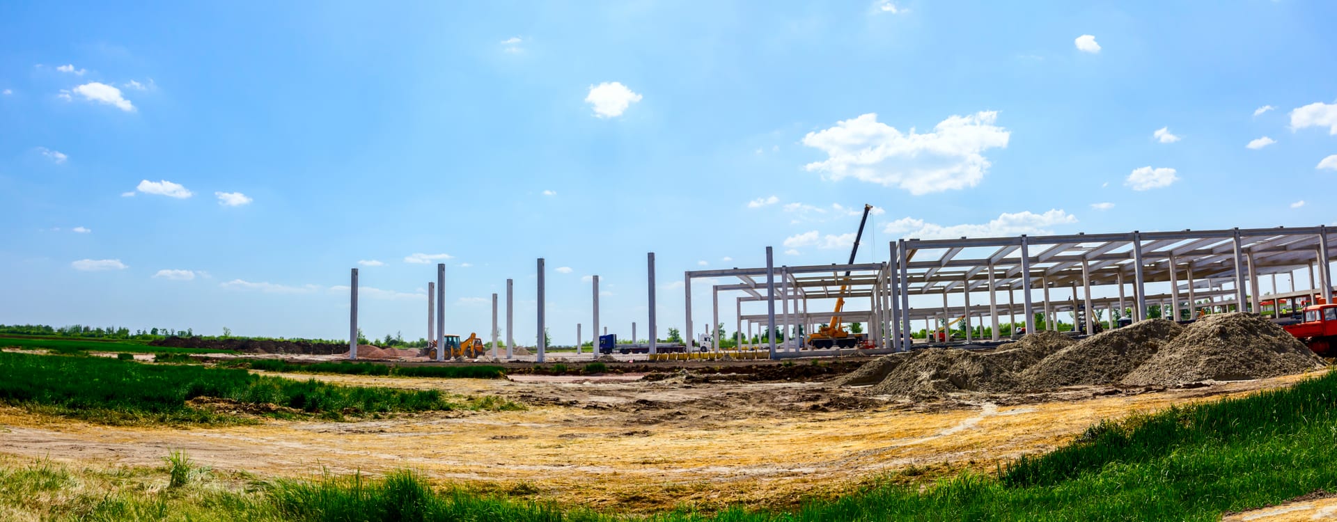 Wide panorama of a new commercial warehouse taking shape on an open site — a mobile crane setting long structural beams across rows of erected columns under a bright blue sky