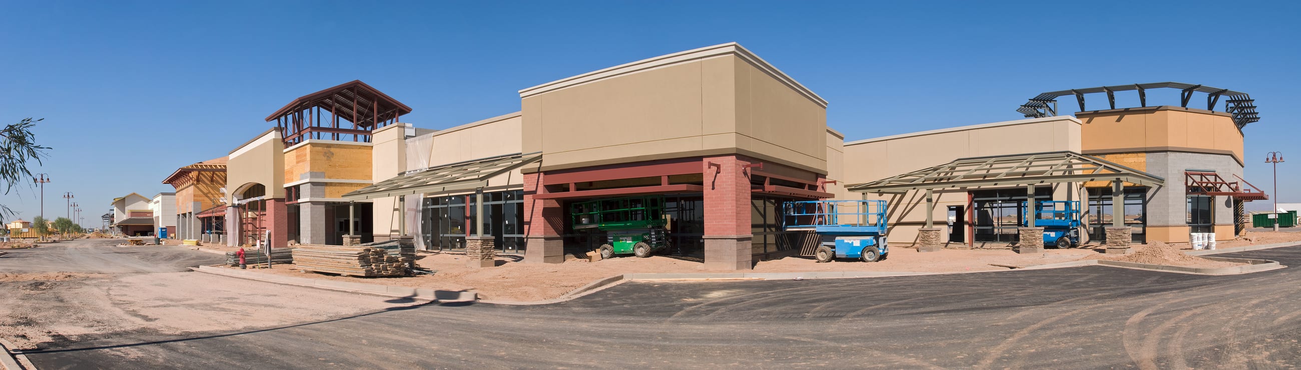 A multi-tenant retail center mid-construction, with stucco and stone storefronts taking shape, steel canopy framing over the walkways, and scissor lifts parked at the building line