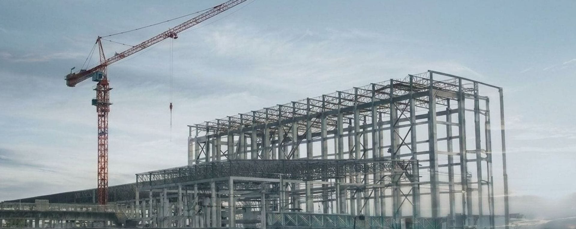 A tower crane standing beside the structural steel frame of a large commercial building under an even, overcast sky