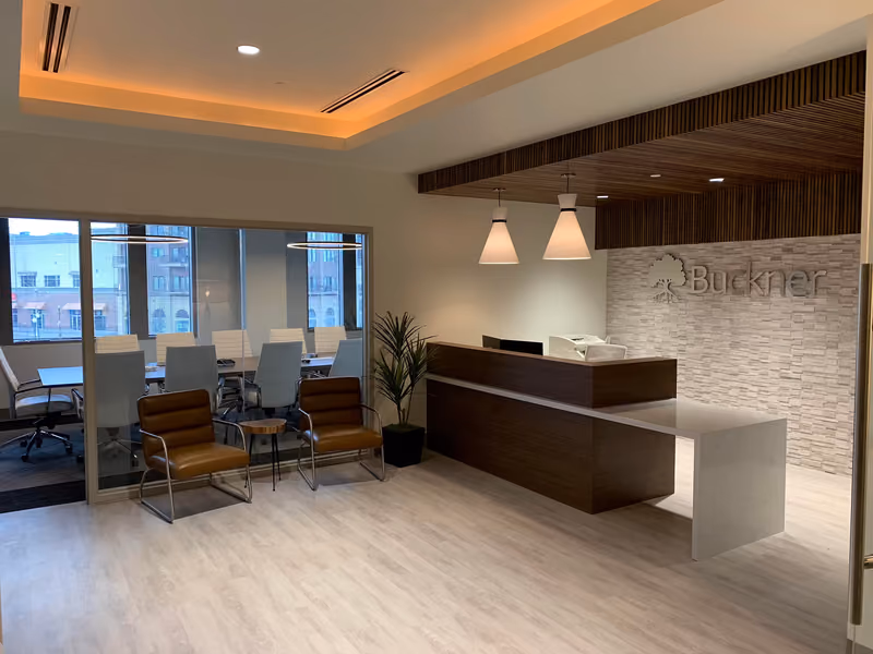 The finished Buckner Insurance reception — a walnut desk under pendant lamps against a stone-faced logo wall, with a glass-walled conference room alongside