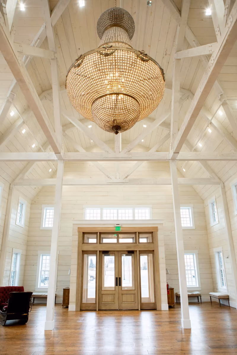 The finished great hall at Walker Farms Event Center — a beaded chandelier hung from an exposed white timber truss ceiling above a hardwood floor, with paned entry doors at the far end