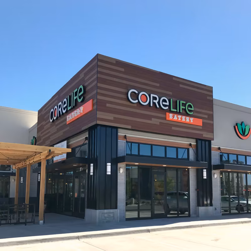 The completed CoreLife Eatery in American Fork — a corner storefront with a wood-slat sign band above black-framed glazing, and a timber pergola over the patio
